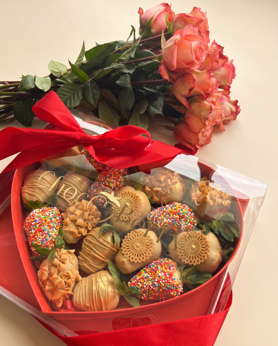 Red heart-shaped gift box with chocolate-covered strawberries and fresh roses above