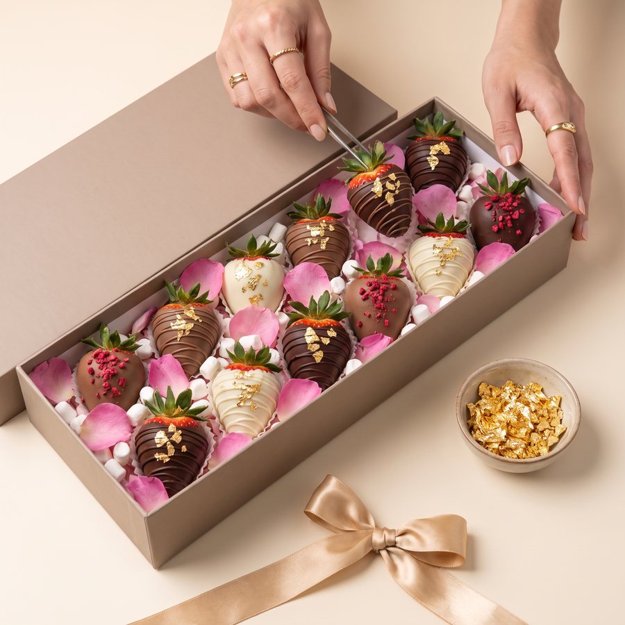 Hands decorating a large open box of chocolate strawberries with gold flakes, rose petals and marshmallows