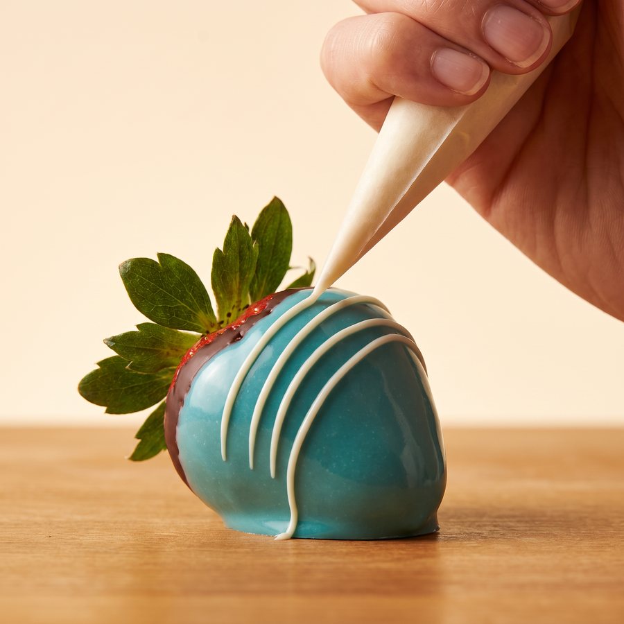 A hand piping thin white chocolate stripes across a turquoise-painted strawberry